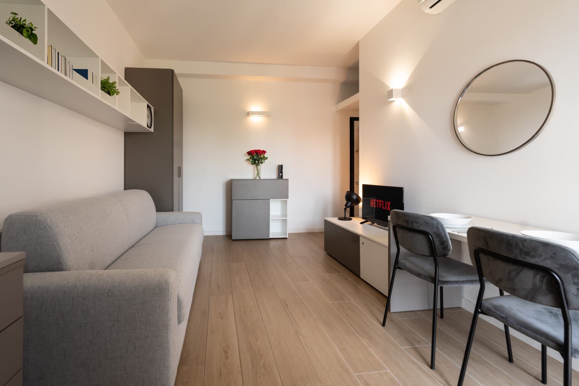 Monolocale allestito per gli ospiti, con divano grigio, mobile TV, specchio tondo e tavolo con sedie